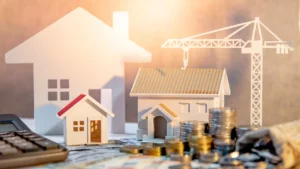 Papier- und Holzmodelle von Häusern mit Baukran und Münzstapeln im Vordergrund – Symbol für Baukosten, Baupreisindex und Immobilienbewertung.